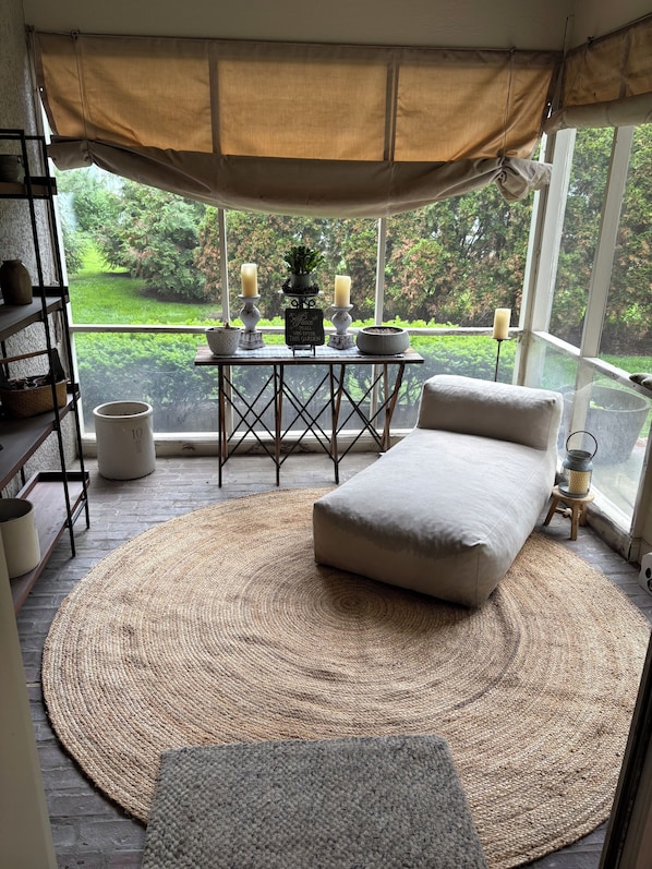 A cozy room with a round woven rug, a cushioned bench, a table with candles, and a view of a lush garden through large windows.
