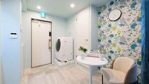 A room with a washing machine, a clock on the wall, a small table with a plant, and a chair.