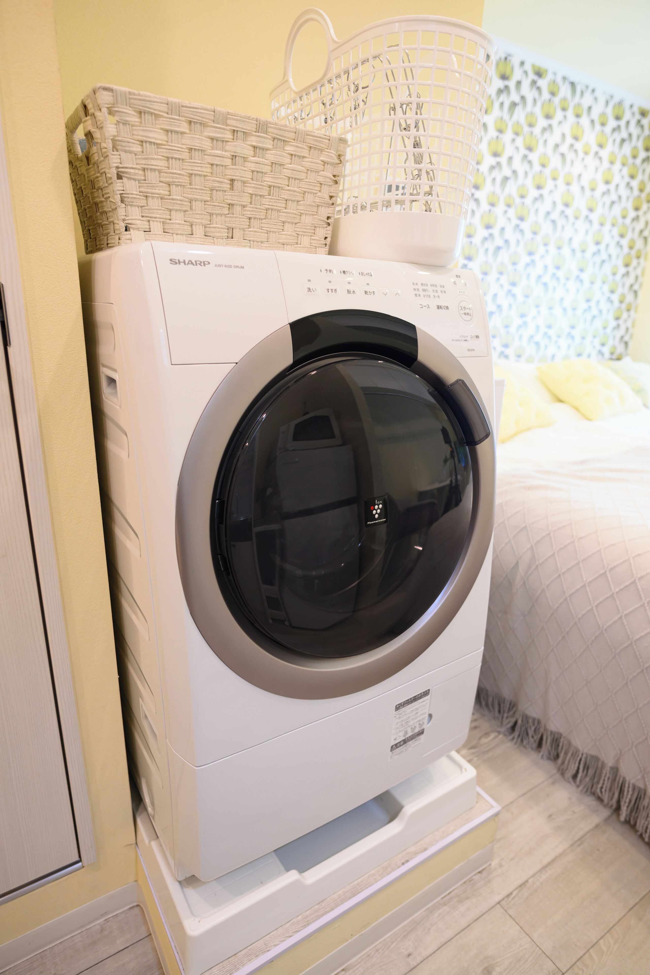 Lavadora de carga frontal con un cesto encima, ubicada en una habitación que tiene una cama y una pared con un diseño llamativo al fondo.