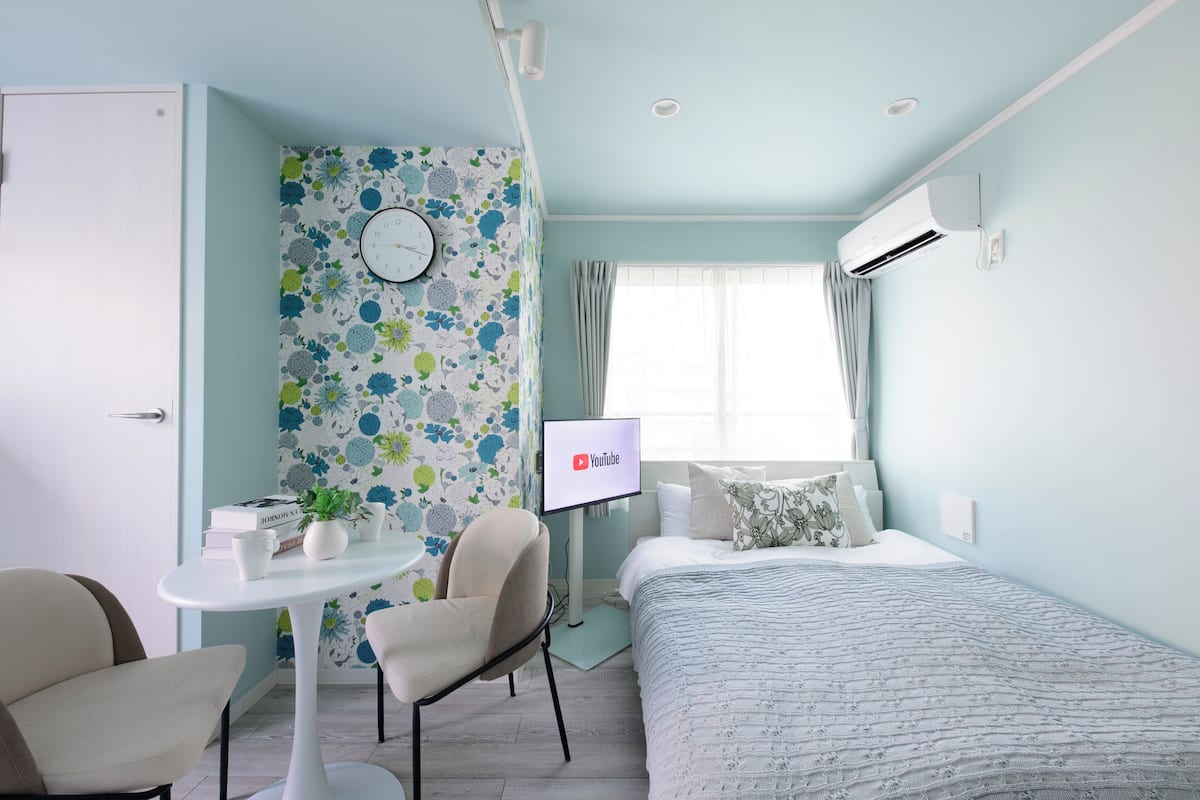 A hotel room with a bed, a desk with a computer, a chair, a clock, and a window with curtains.