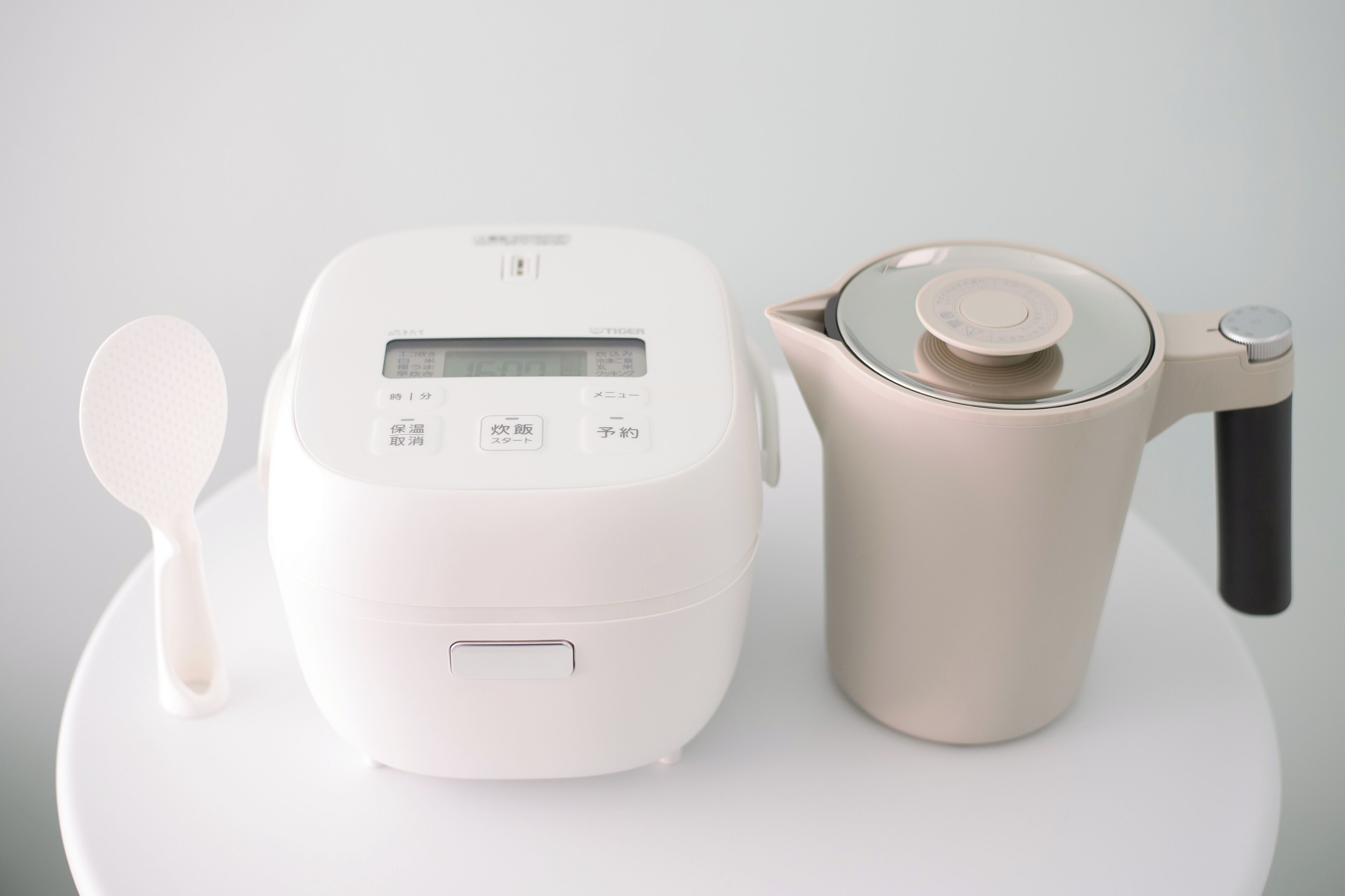 A white rice cooker with a digital display and buttons, a beige pot with a lid, and a white spoon on a white surface.