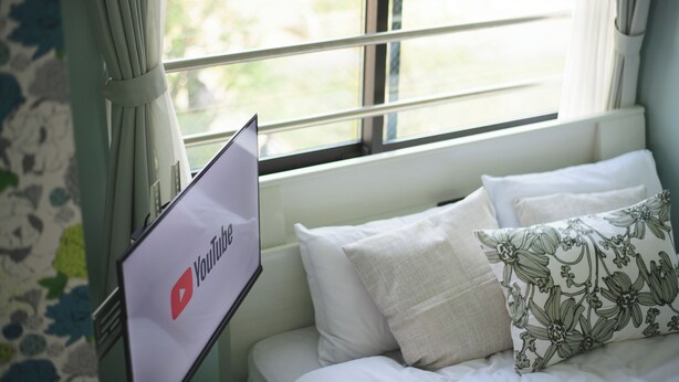 A bed with white bedding and a floral pillow, a laptop displaying the YouTube logo, and a window with curtains.