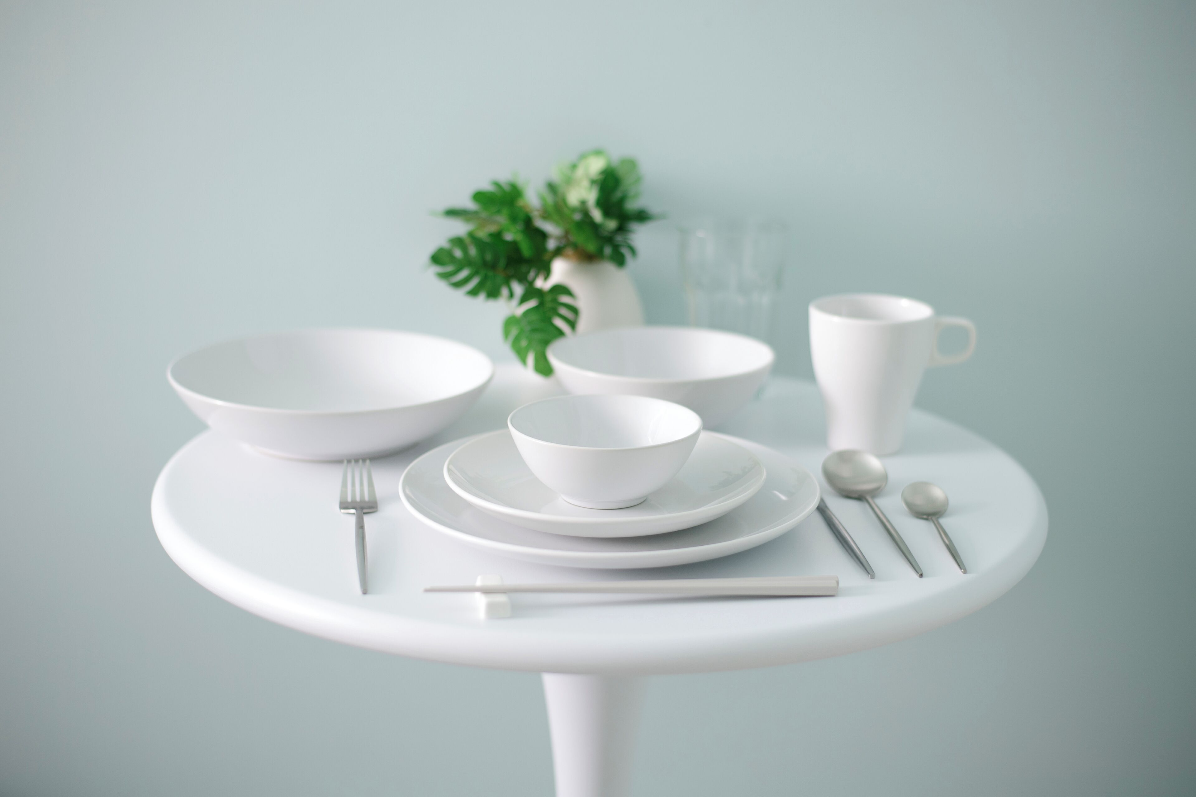 A minimalist dining setup with white bowls, plates, a cup, and cutlery on a white table.
