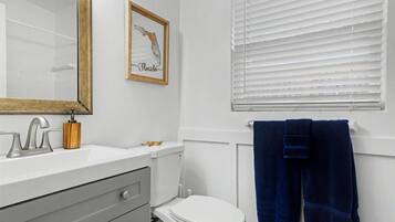 Un baño con inodoro blanco, lavamanos con espejo y ventana con persianas.