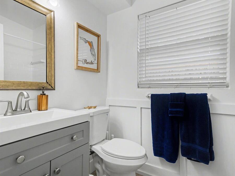 Un baño con inodoro blanco, lavamanos con espejo y ventana con persianas.