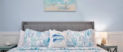 Un dormitorio con una cama con colcha azul y blanca de motivos florales, un cabecero tapizado en gris y un cuadro con una playa sobre la cama.