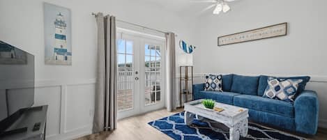 A living room with a blue sofa, a coffee table, a ceiling fan, and a door leading to a balcony.