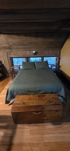A wooden cabin bedroom with a bed, bench, and windows overlooking a forest.