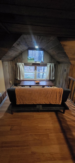 A cozy attic room with a wooden ceiling, a large window with curtains, a wooden floor, and a sofa with a blanket.