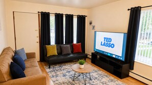 A living room with a sofa, a coffee table, a TV displaying 'TED LASSO,' and a window with curtains. - The Arbor's Haven - Cozy, Stylish Retreat Steps from Downtown Ann Arbor (Ann Arbor)