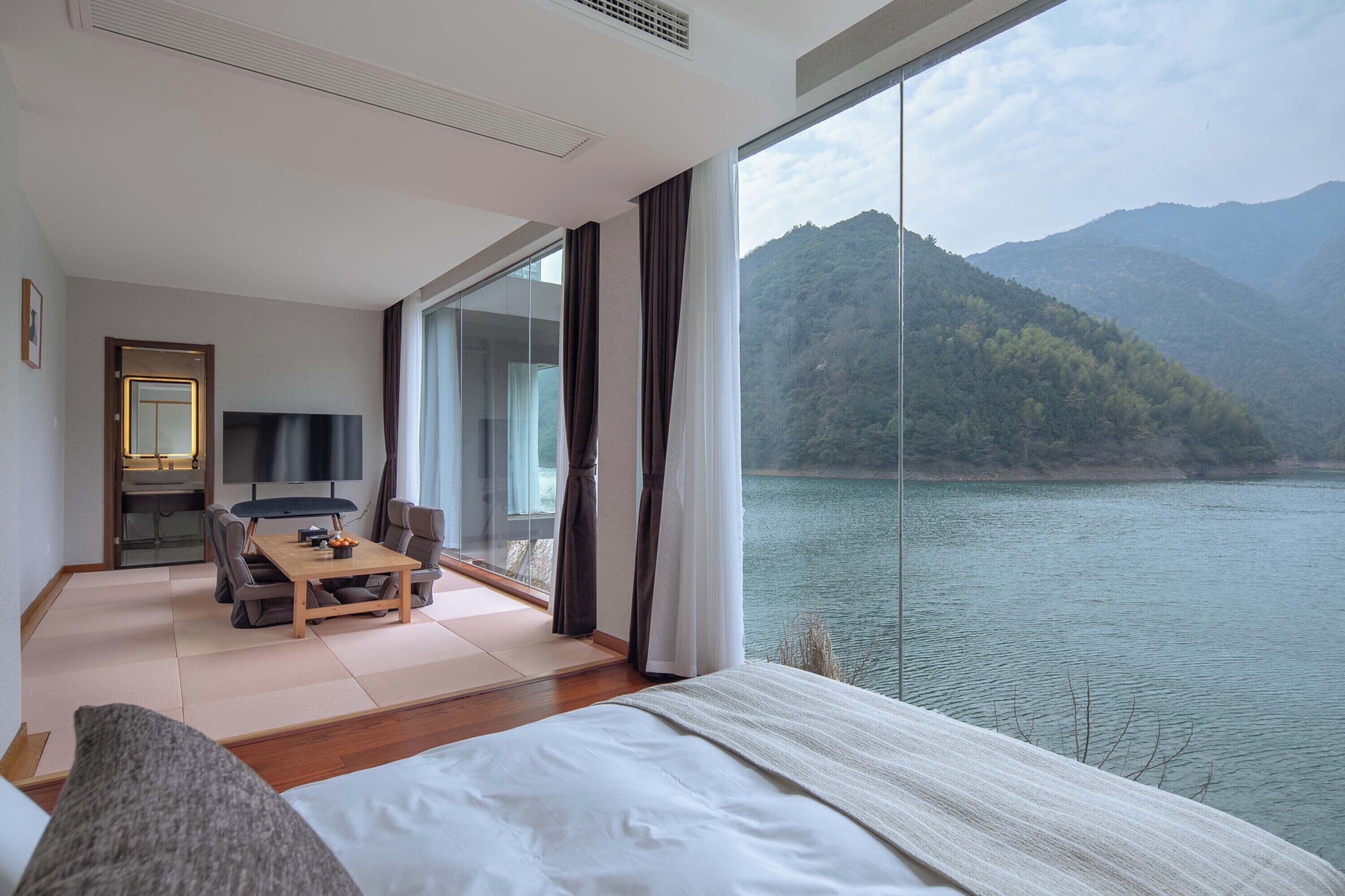 A hotel room with a large window overlooking a lake and mountains, a bed, a dining area with a table and chairs, and a TV.
