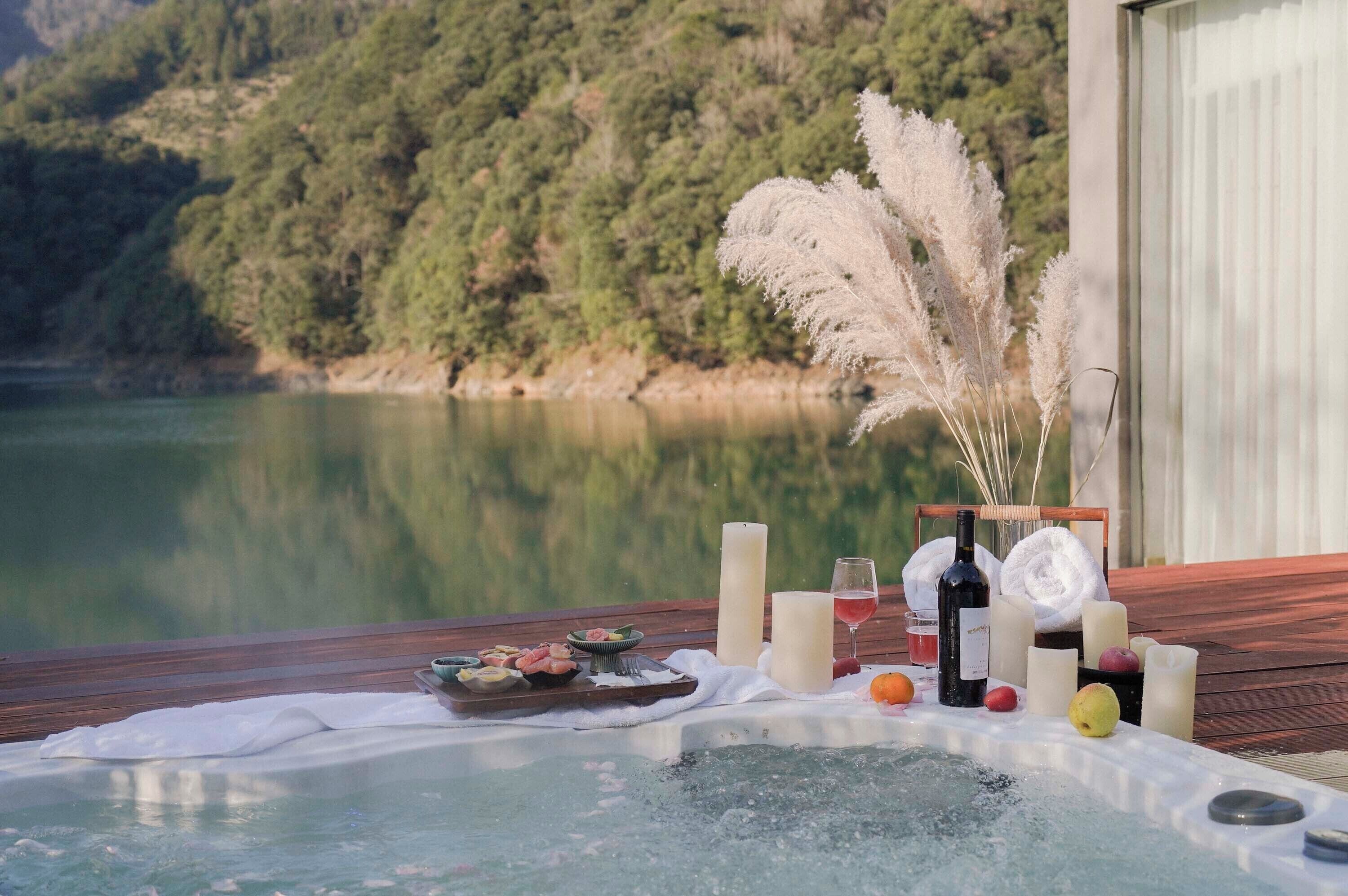 Een hottub met een eettafel, kaarsen en een fles wijn, met uitzicht op een rustig meer en beboste heuvels.