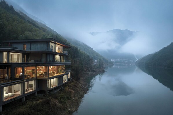 A modern multi-story building with large windows overlooking a calm river, surrounded by forested hills. - LOST LODGE Shendu Wubei Lin'an (Hangzhou)