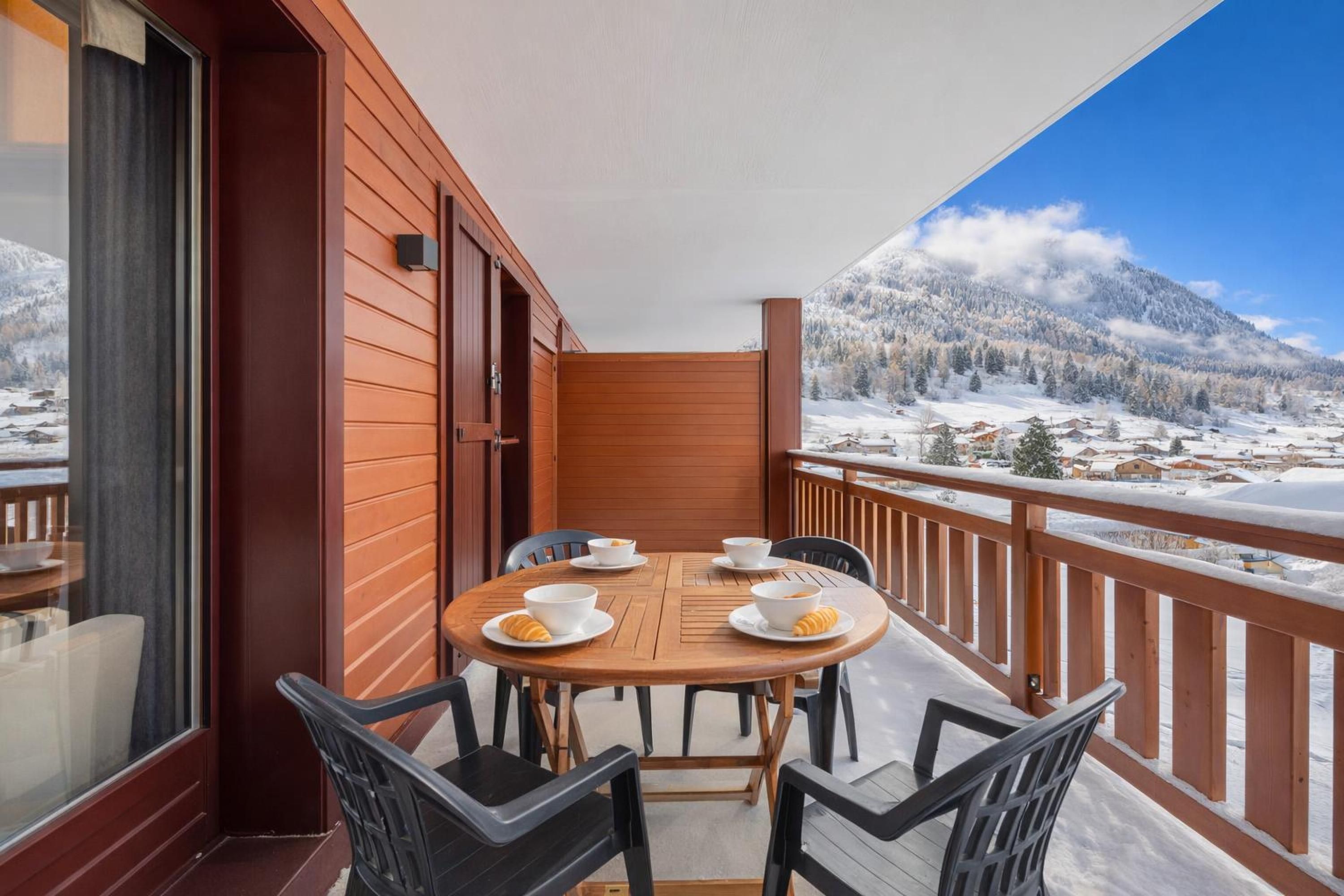 En balkon med et træbord og stole, der ud til et sneklædt bjerglandskab.