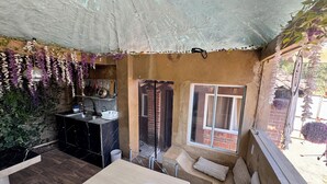 A cozy outdoor seating area with a wooden table and cushioned benches, a kitchen area with hanging plants, and a view of the street through a glass door. - RUSTIC CABIN IN THE CENTER OF TEPOZTLAN (Tepoztlán)