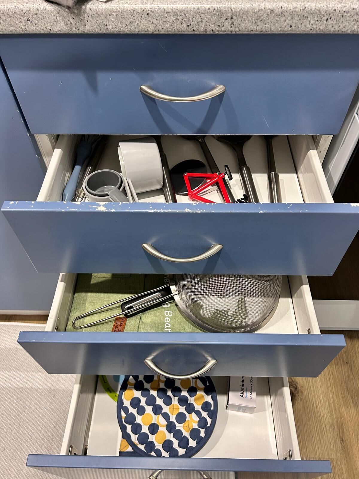 A drawer with multiple compartments, containing kitchen utensils, a strainer, and a patterned dish.