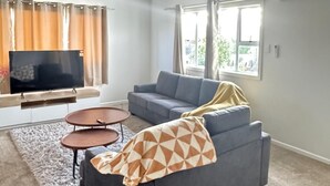 Ett modernt vardagsrum med en grå soffa, två små, runda bord, en platt-TV och ett stort fönster med gardiner.