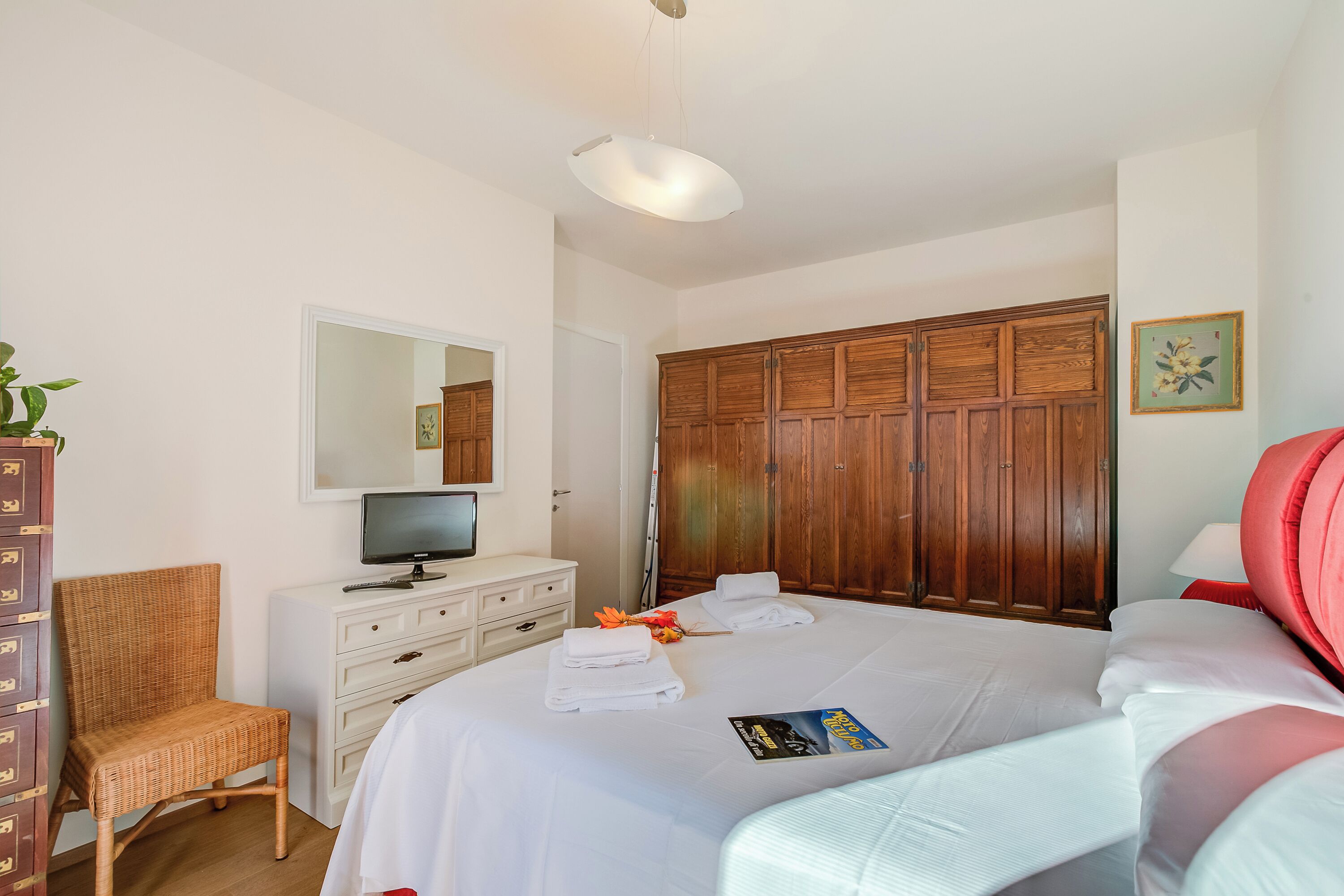 A bedroom with a bed, a television on a dresser, a wardrobe, a chair, and a bedside table.