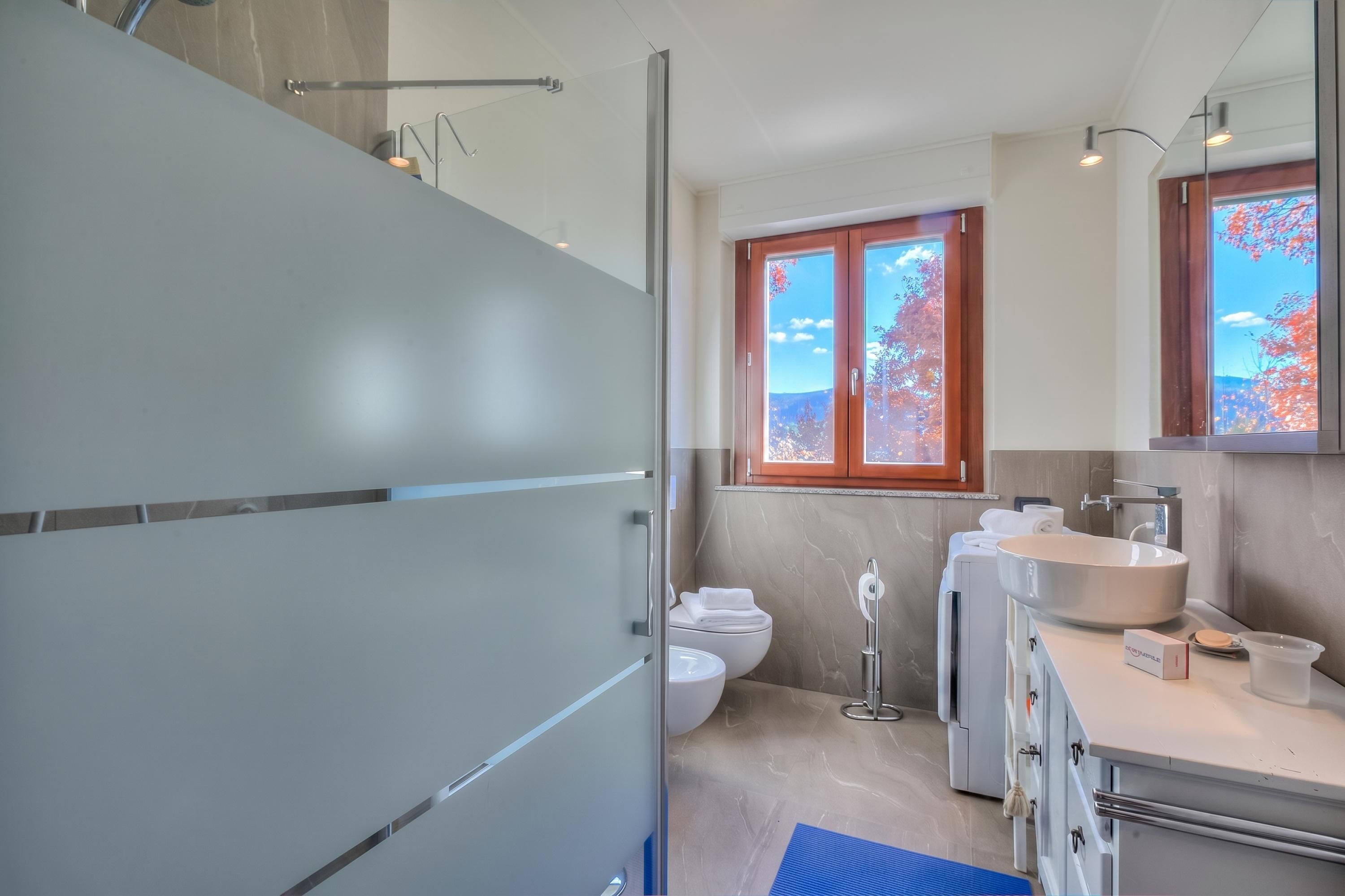 A modern bathroom with a glass shower enclosure, a toilet, a sink with a mirror, and a window with a view of trees.