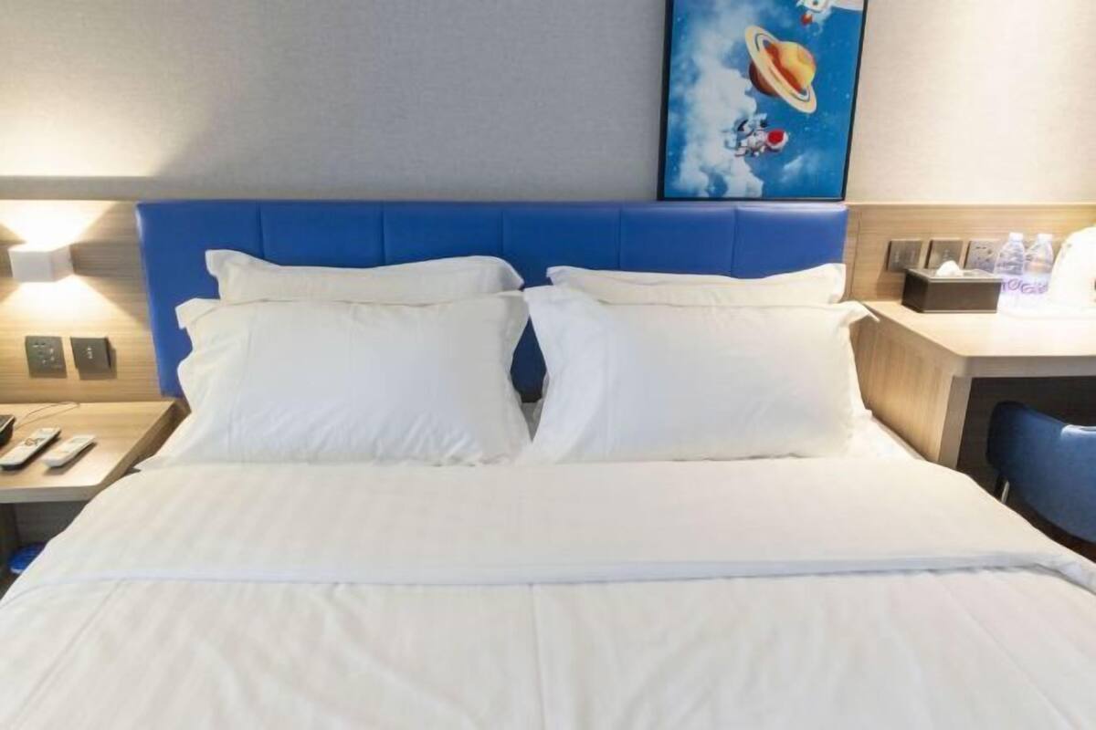 A hotel room with a blue headboard, white bedding, a bedside table with a lamp, and a painting on the wall.