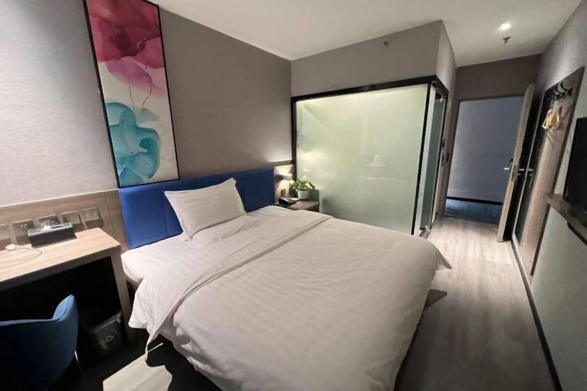 A hotel room with a bed, a desk, a chair, a nightstand, and a large window.