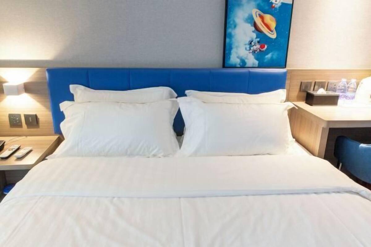 A hotel room with a blue headboard, white bedding, a desk with a chair, and a wall-mounted painting.