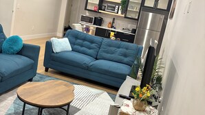 A modern living room with a blue sofa set, a wooden coffee table, and a kitchen area with stainless steel appliances. - The Cozy Haven (Halifax)