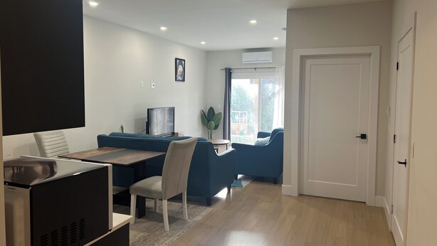 A modern living room with a blue sofa, a dining table, and a TV. There is a door leading to another room and a window with a view of the outside. - The Cozy Haven (Halifax)