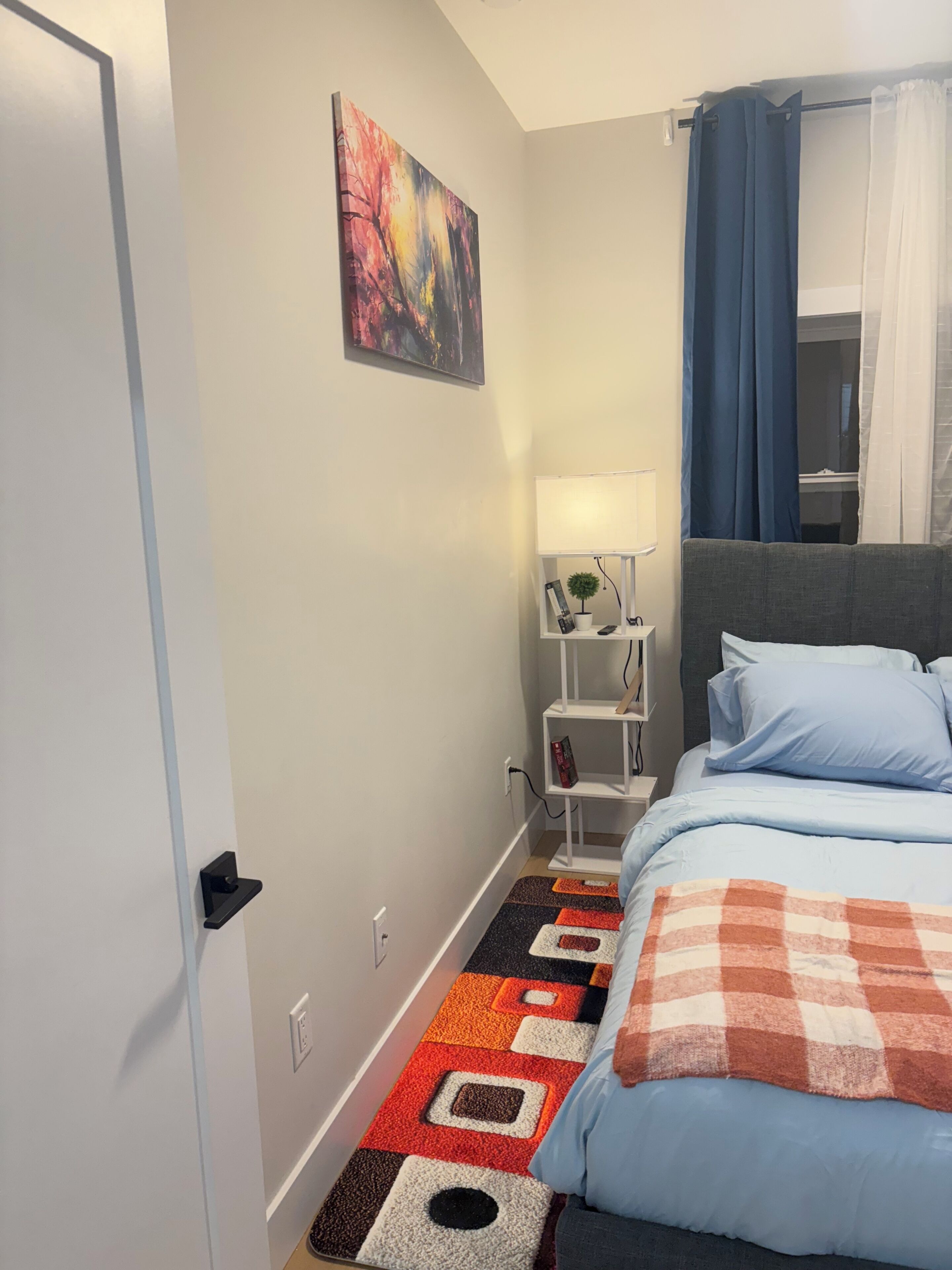 A bedroom with a bed, a nightstand, a lamp, a painting, and a window with curtains.
