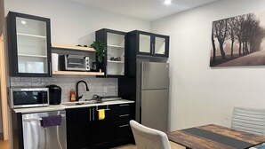 A modern kitchen with black cabinets, a stainless steel refrigerator, a microwave, and a sink. There is a dining table with a chair and a blue sofa. - The Cozy Haven (Halifax)
