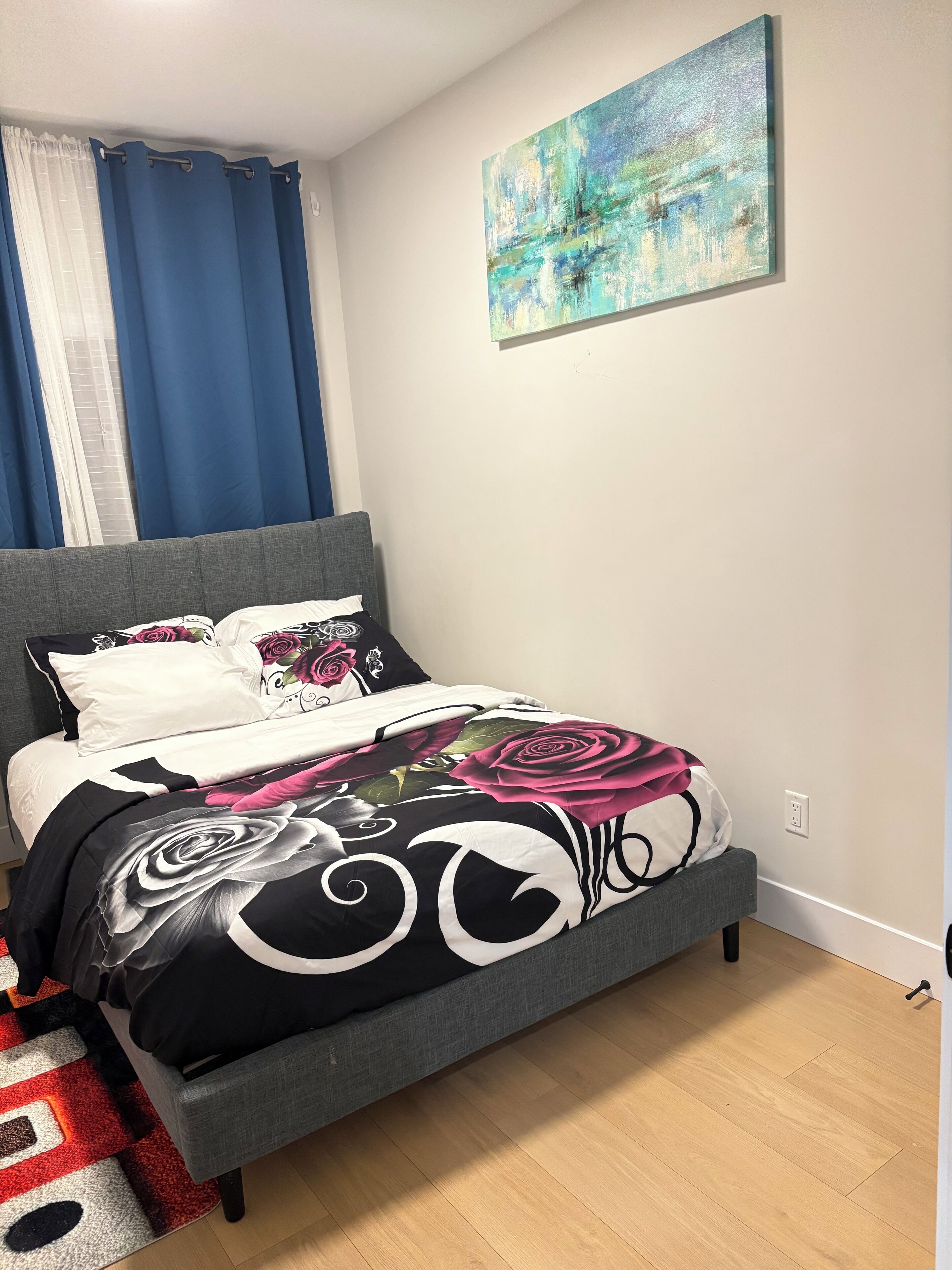 A bedroom with a grey upholstered bed, a colorful floral bedspread, and a blue curtain.