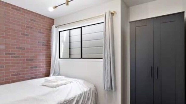A bedroom with a brick wall, a bed with a white cover, a window with curtains, and a dark wardrobe. - Blue&Gold Art Deco 2BR1BA Loft close to Genentech (South San Francisco)