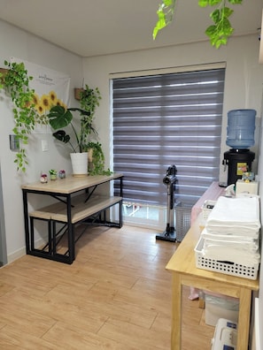 A room with a wooden floor, a table with a water dispenser, a bench, and a large window with blinds. - Incheon Airport ANN Guesthouse (Incheon)