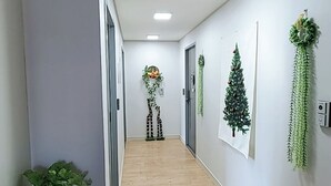 A hallway with wooden flooring, white walls, and ceiling lights. There are decorative plants and a small Christmas tree on the walls. - Incheon Airport ANN Guesthouse (Incheon)