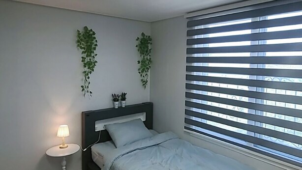 A bedroom with a bed, a bedside table with a lamp, a ceiling light, and a window with blinds. - Incheon Airport ANN Guesthouse (Incheon)