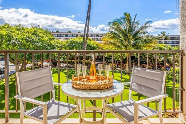 A balcony with two white chairs, a round table, and a decorative centerpiece. - Partial oceanview 1 bedroom condo, AC, across from beach, near shops & dining (Kihei)