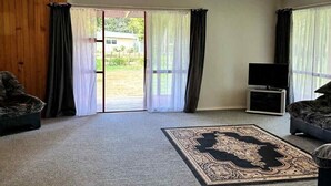 A spacious living room with a large sliding glass door leading to a backyard, a flat-screen TV, and a patterned rug. - Paradise At The Beach (Coromandel)