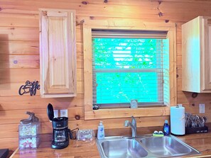 Un interior de madera con ventana, cafetera, lavamanos y una estantería con utensilios de cocina.