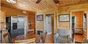Interior de cabaña con revestimiento de madera, zona de comedor con mesa y sillas, refrigerador, lavamanos y un sillón estampado.
