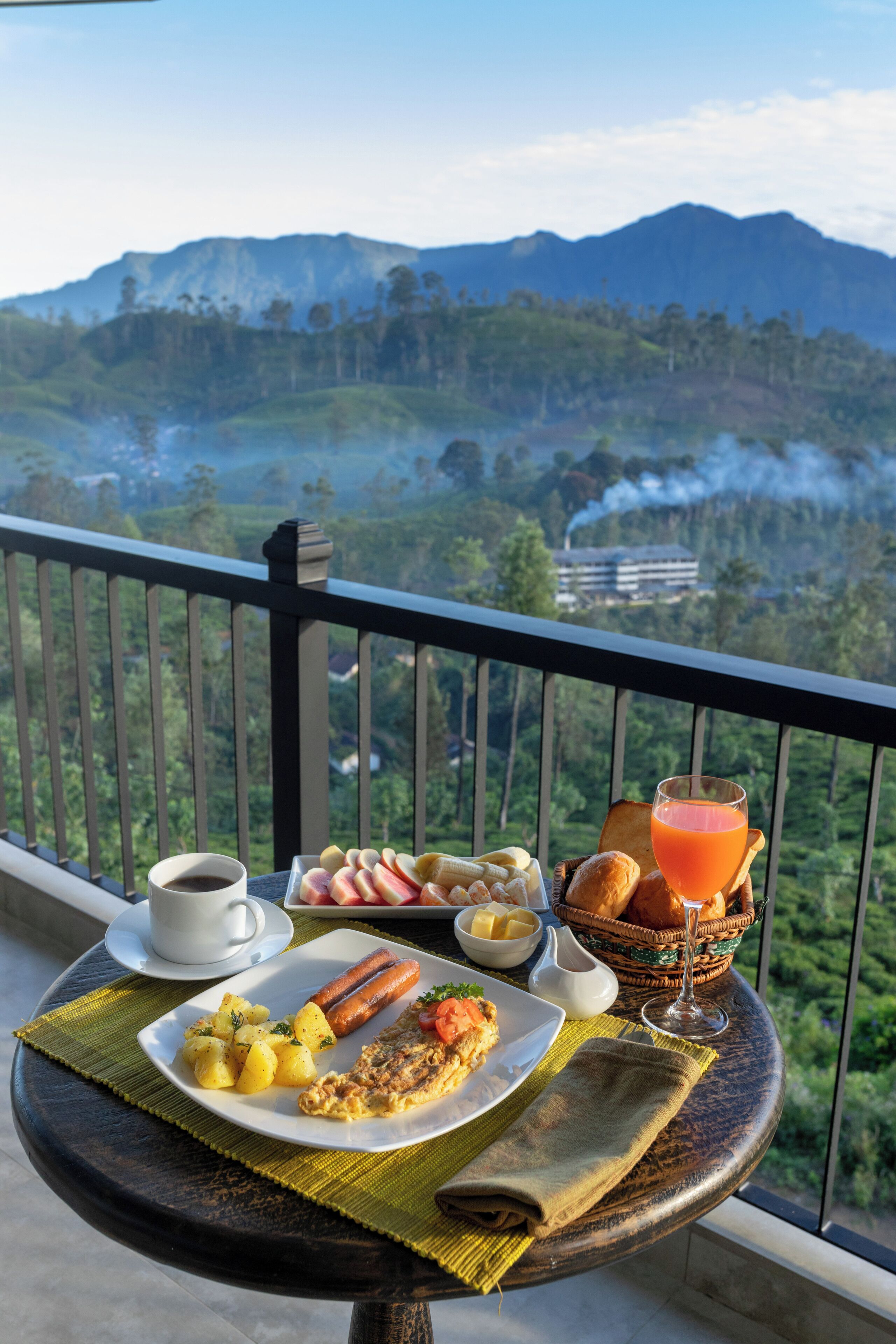 Een balkon met een tafel gedekt voor het ontbijt, inclusief eieren, worst, aardappelen, toast en een glas sinaasappelsap, met uitzicht op een bergachtig landschap.