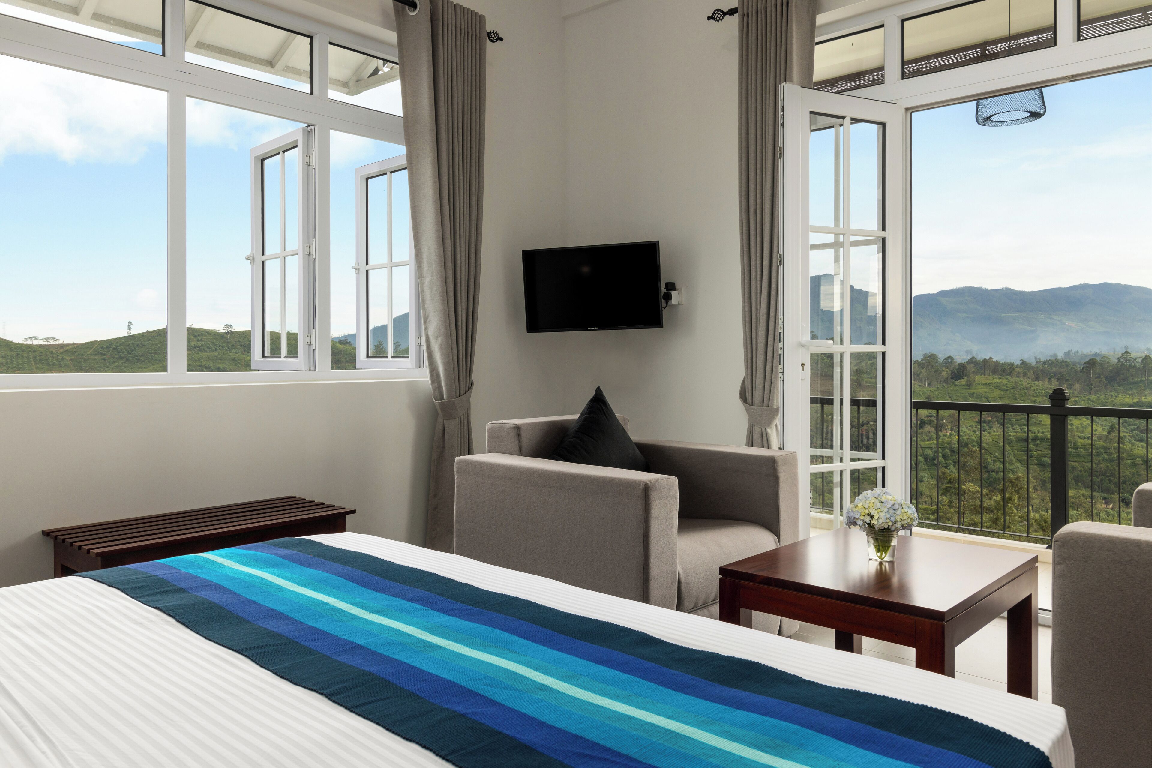 A hotel room with a bed, a TV mounted on the wall, a chair, a small table with a vase of flowers, and large windows offering a view of the mountains.