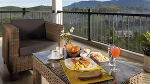 A balcony with wicker furniture, a dining table with breakfast items, and a scenic mountain view. - Ruvinya Hills Luxury Bungalow (Lindula)
