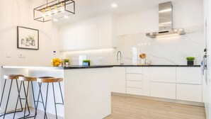 Eine moderne Küche mit weißen Schränken, einer schwarzen Arbeitsplatte und einer Edelstahl-Dunstabzugshaube. Drei Barhocker mit Holzsitzen und schwarzen Metallbeinen sind vorhanden.