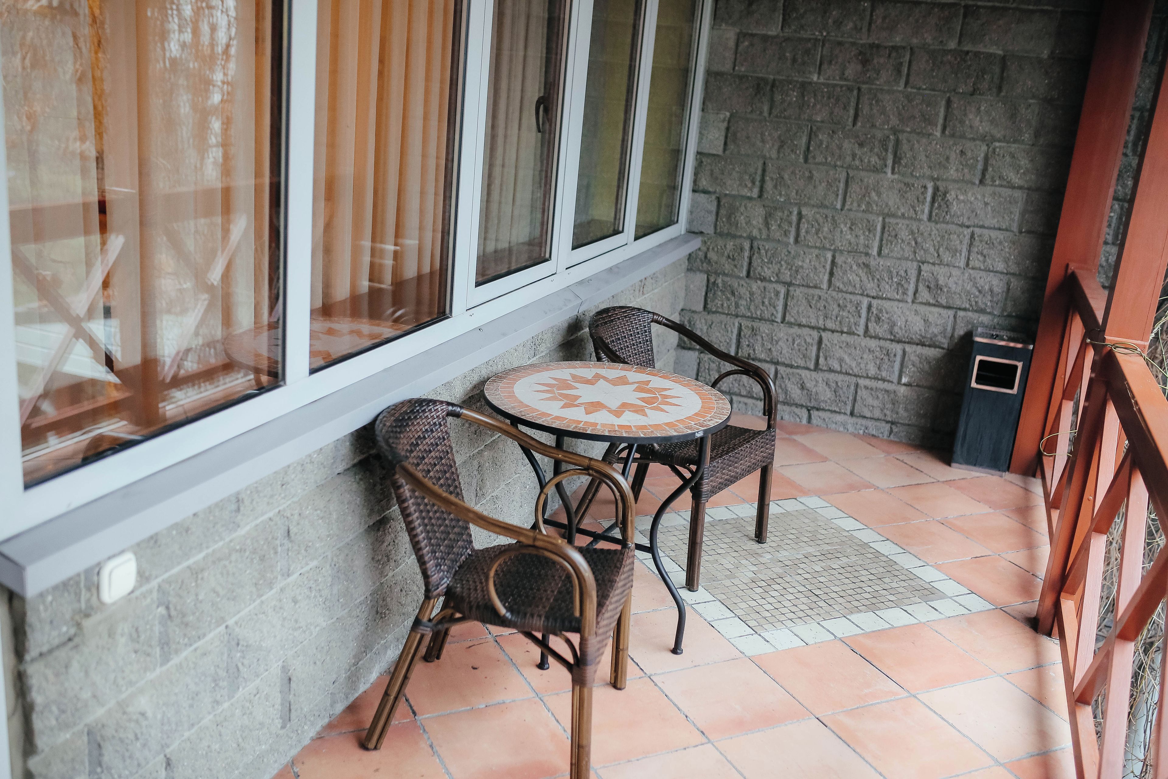 A small outdoor seating area with two wicker chairs and a round table, a window with curtains, and a brick wall.