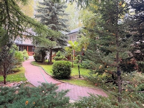 A garden pathway leading to a building with a red roof, surrounded by various trees and shrubs.