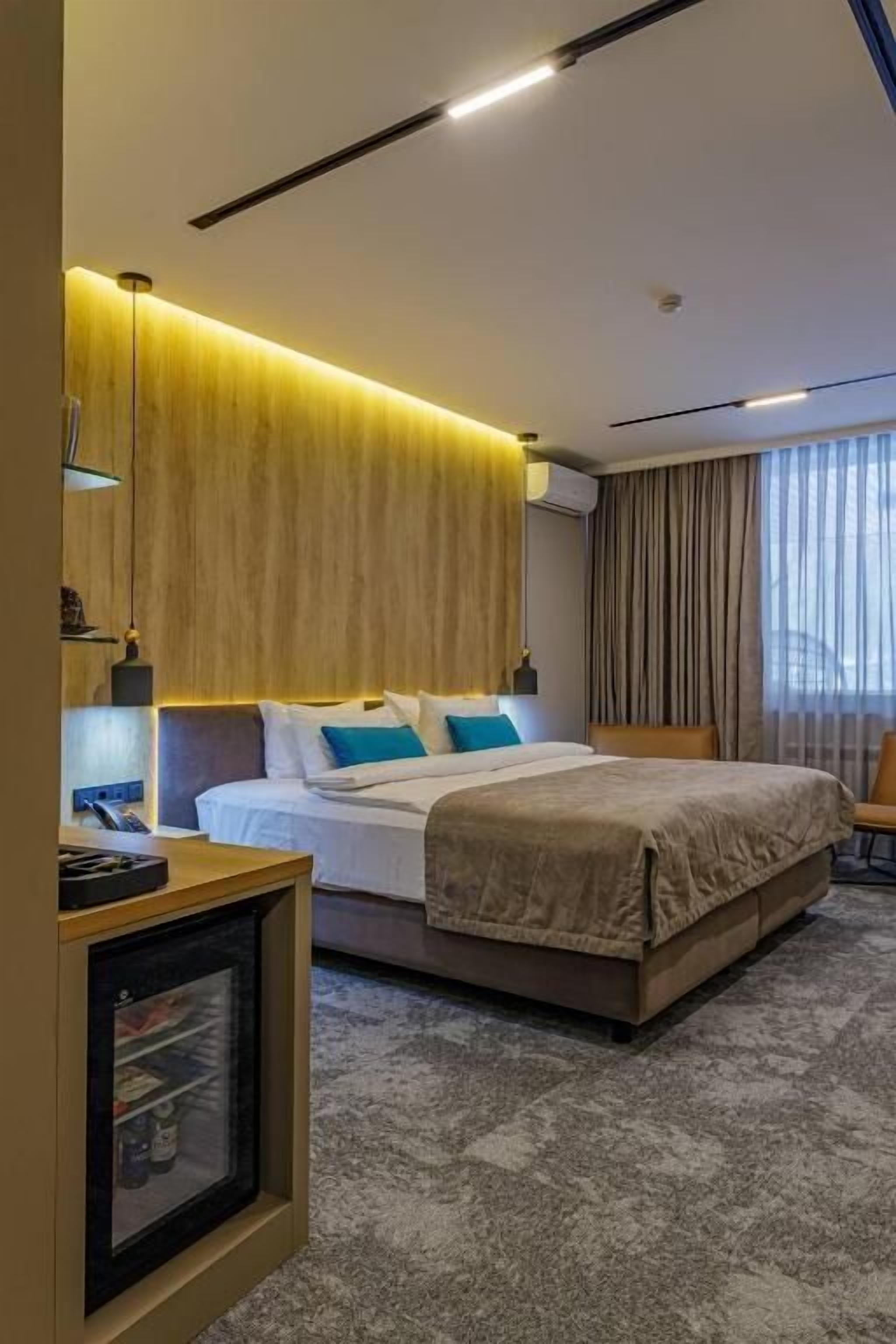 A modern hotel room with a large bed, a bedside table, a small fridge, and a view of the outdoors through a window.