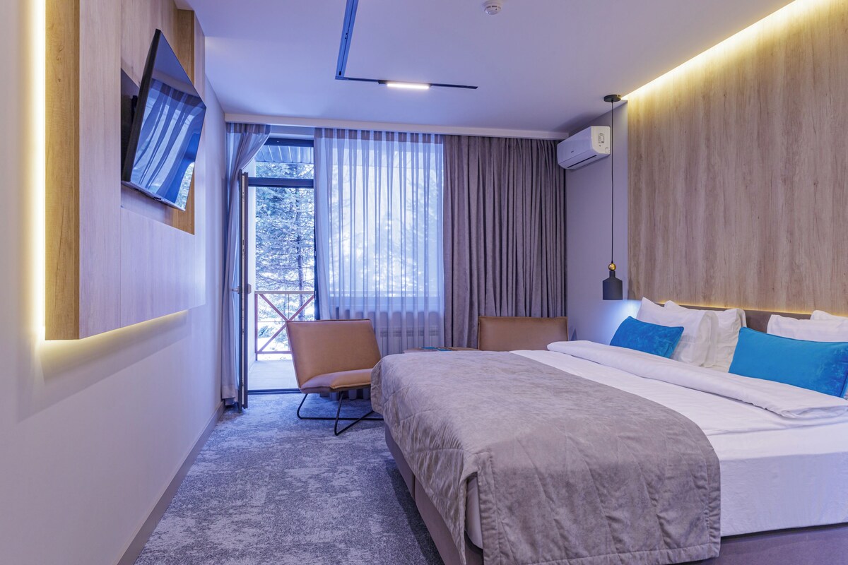 Una habitación de hotel moderna con una cama, una silla, un televisor montado en la pared y un ventanal con cortinas.
