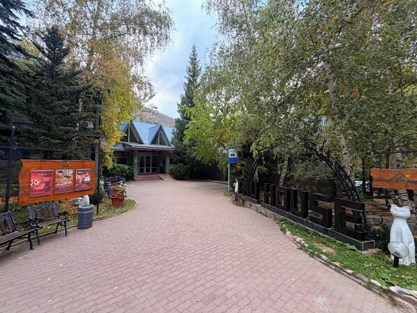 A paved pathway leading to a building with a sloped roof, surrounded by trees and benches. - TAU-House (Almaty)