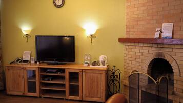 Una sala de estar con televisor de pantalla plana, mueble bajo de madera, dos lámparas de pared, chimenea de ladrillo y un reloj en la pared.