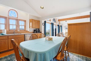 Un coin repas avec une table dressée pour quatre personnes, des chaises en bois, un espace cuisine avec des placards en bois, et une vue par la fenêtre.